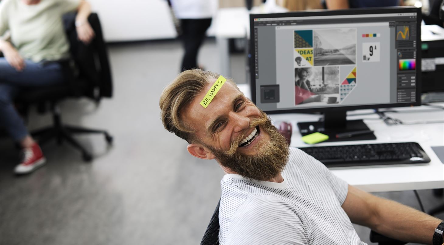 En glad mand sidder ved et skrivebrod bag en computer med en seddel i panden, hvorp der stt "be happy".