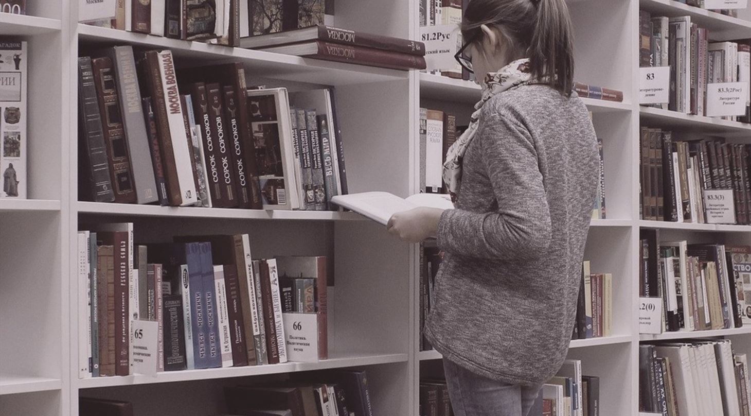 Et billede af en person, der står og kigger i en bog på et bibliotek.