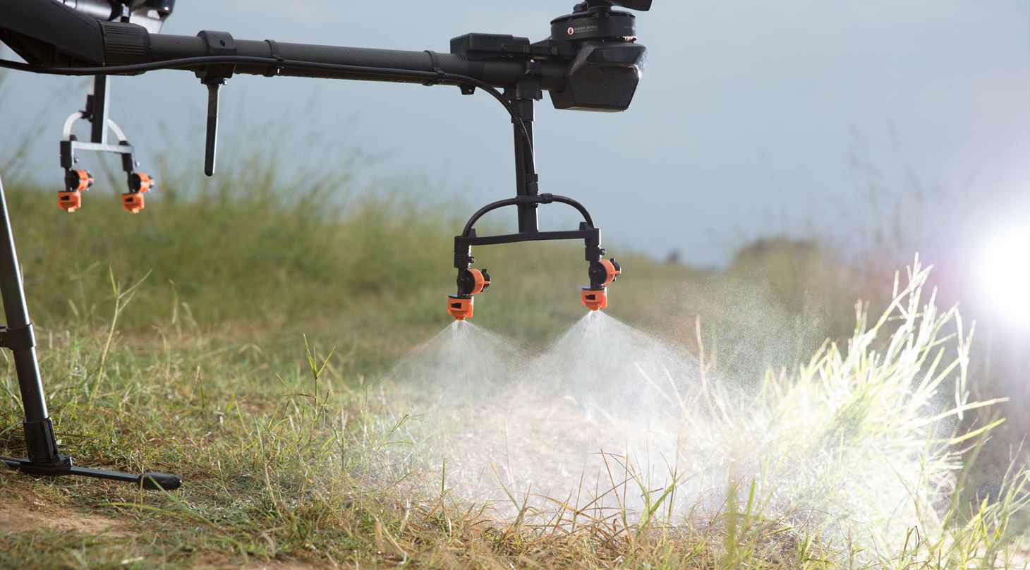 På billedet ser man en drone, der har et værktøj, en sprøjte koblet på, som er ved at sprøjte på en mark.