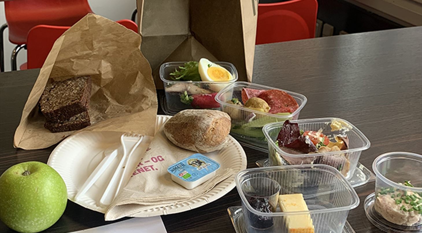 Billedet viser en frokostpakke to-go sat op, så man kan se de forskellige elementer frokostpakken indeholder, dvs. forskellige retter i plastbokse samt brød i papirpose