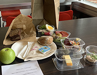 Billedet viser en frokostpakke to-go sat op, så man kan se de forskellige elementer frokostpakken indeholder, dvs. forskellige retter i plastbokse samt brød i papirpose