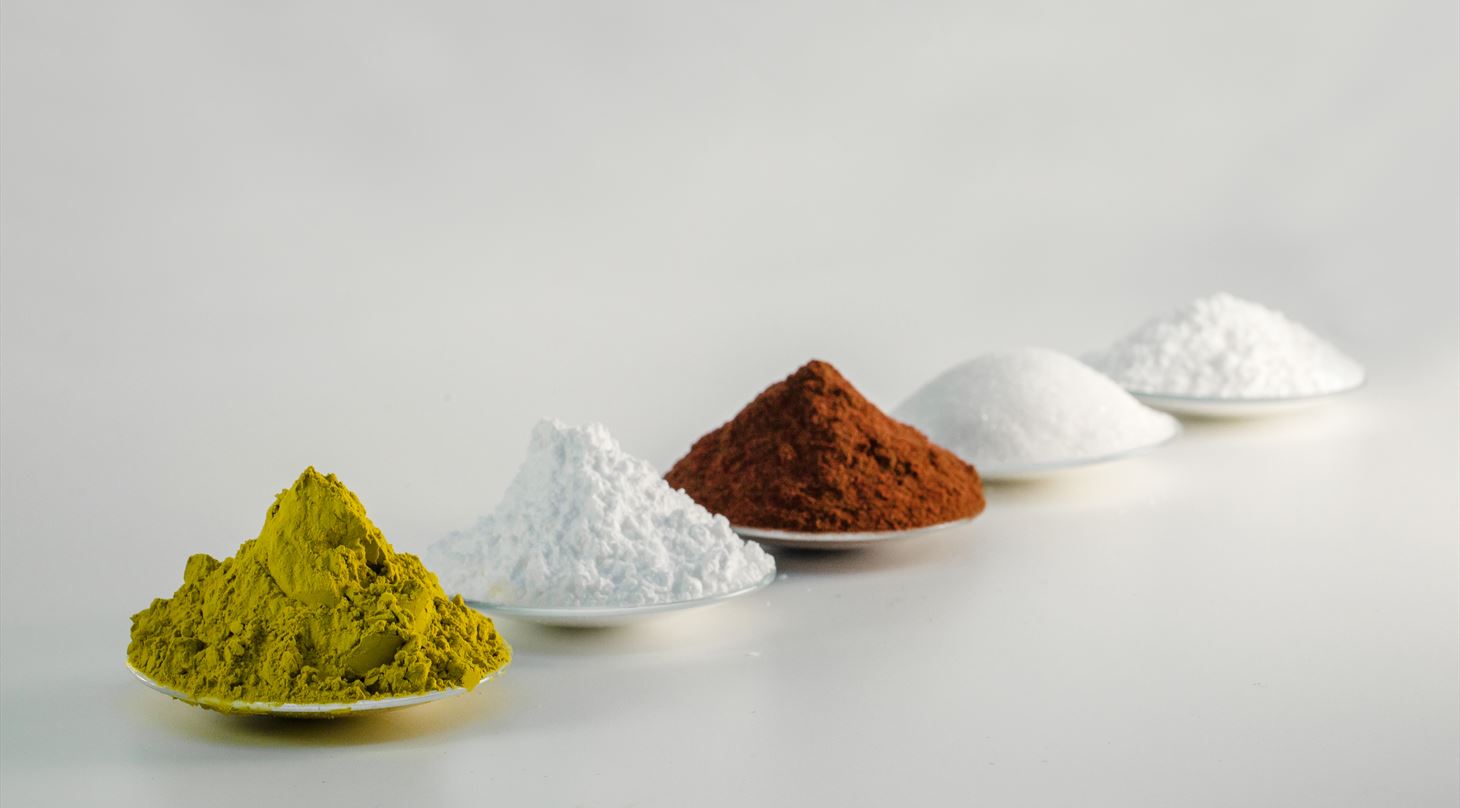 Billedet viser fem forskellige pulvere i forskellige farver og formalingsgrader. Først et gulligt fint pulver med høj top, dernæst et fint hvidt pulver også med høj top, så et grovere orange pulver, et groft hvidt pulver med rund top og sidst hvidt pulver