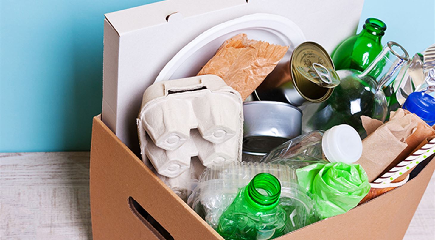 Billedet viser en papkasse fyldt med forskellige slags affaldsemballager, bl.a. dser, glasflasker, plastflasker, karton og papir
