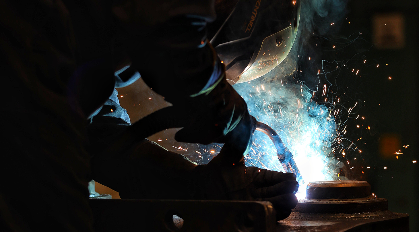 En svejser i beskyttelsesdragt og hjelm arbejder intenst med en metalgenstand, mens gnisterne flyver og skaber et dramatisk lysspil. De blå og orange gnister står i kontrast til den mørke baggrund, hvilket fremhæver svejseprocessens intensitet.