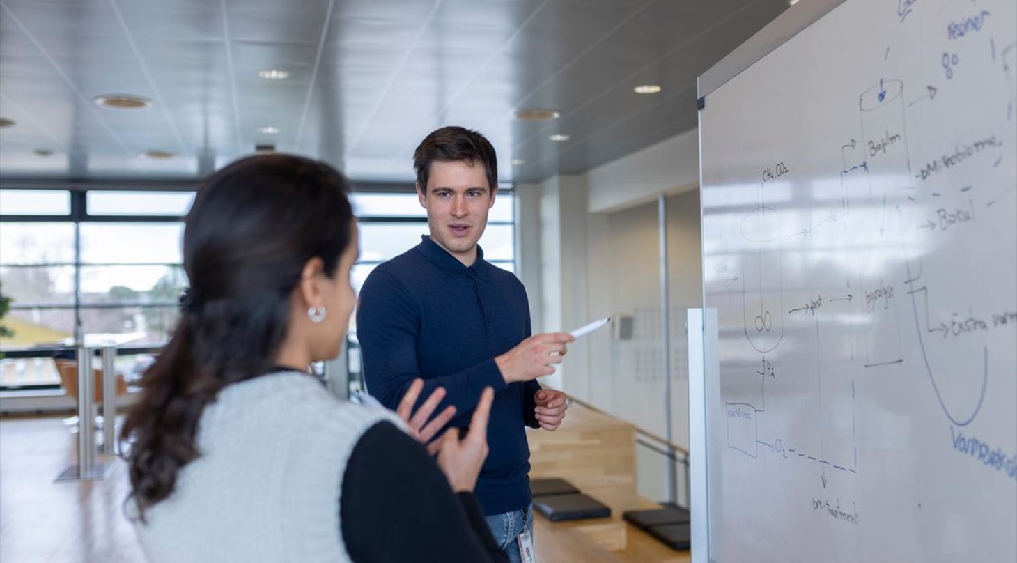 To personer str ved et whiteboard i et lyst kontormilj og diskuterer en teknisk tegning eller diagram, mens den ene peger p tavlen med en tusch.