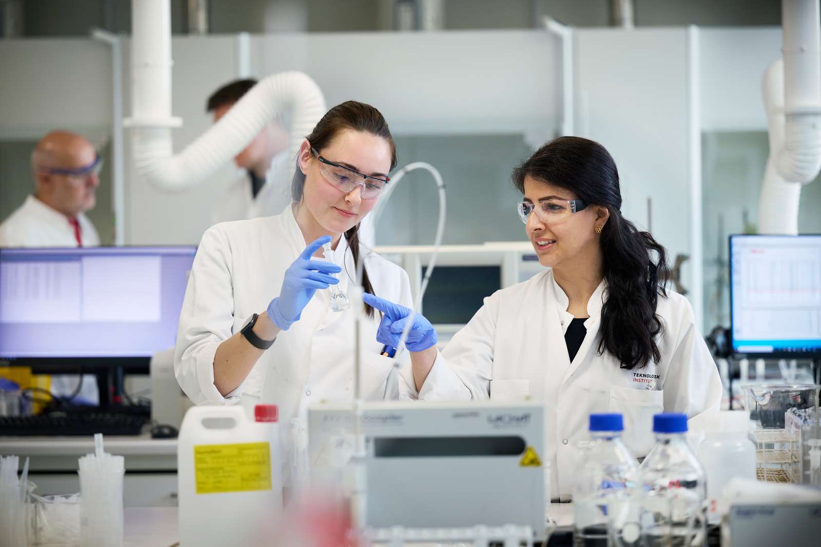 2 kvinder st�r i laboratoriet i kitler og briller og kigger p� analyser