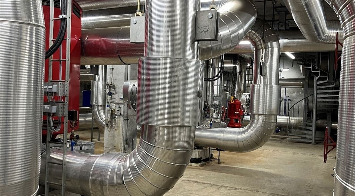 Industrielt teknikrum med store isolerede r�r, ventiler og metaltrappe.