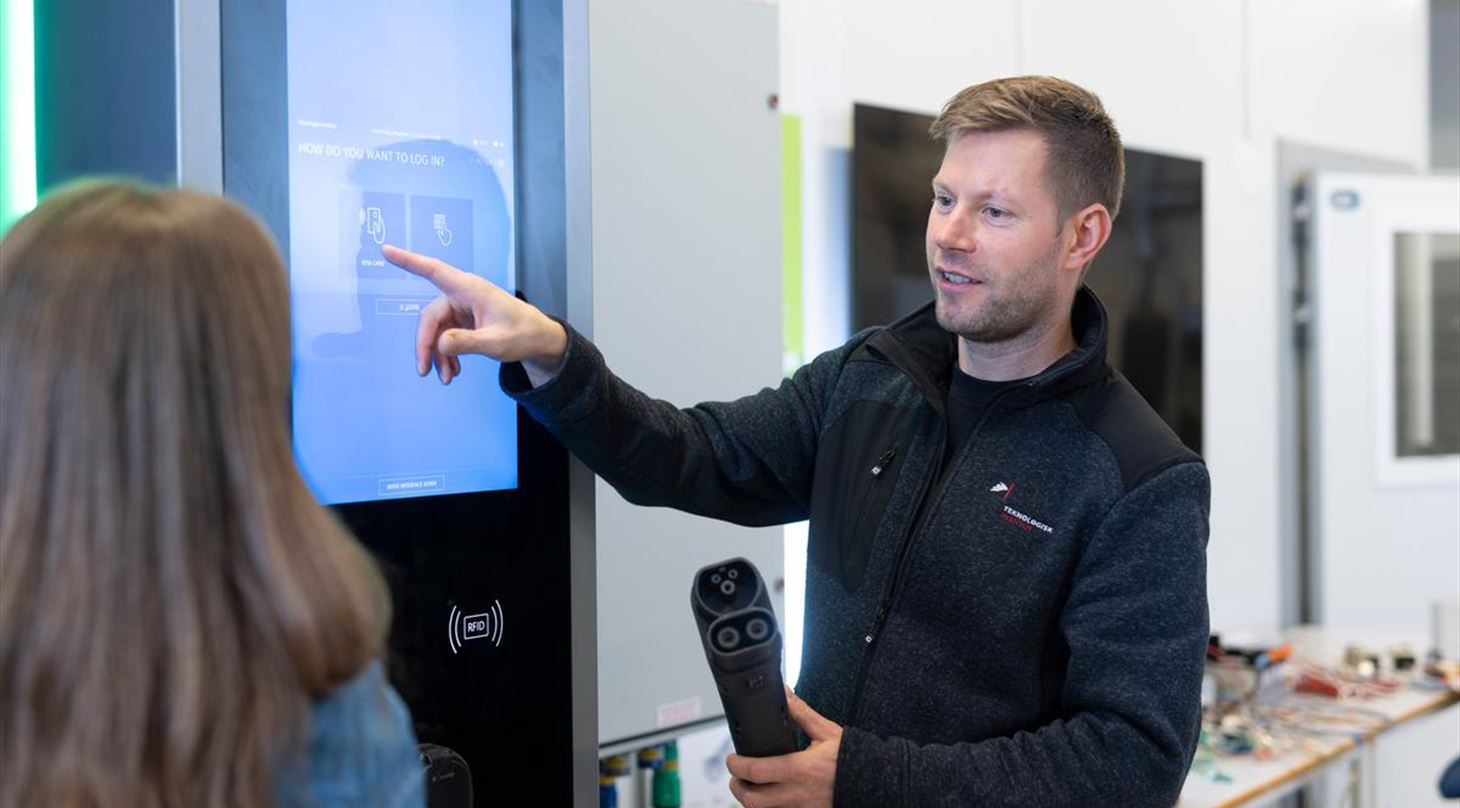 Mandlig medarbejder fra Teknologisk Institut holder et ladestik til en elbil i hånden og peger på en digital ladestander med teksten 'How do you want to log in?', mens han taler med en kvinde i et laboratorium.