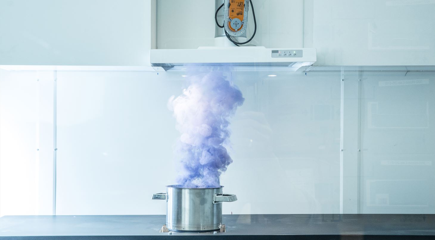 En gryde i stål på en kogeplade, hvorfra en tæt sky af lilla-blålig røg stiger direkte op mod en emhætte i et hvidt køkken- eller laboratoriemiljø.
Gryde på en kogeplade i et laboratorium, med en sky af lilla-blålig røg stiger direkte op mod en emhætte